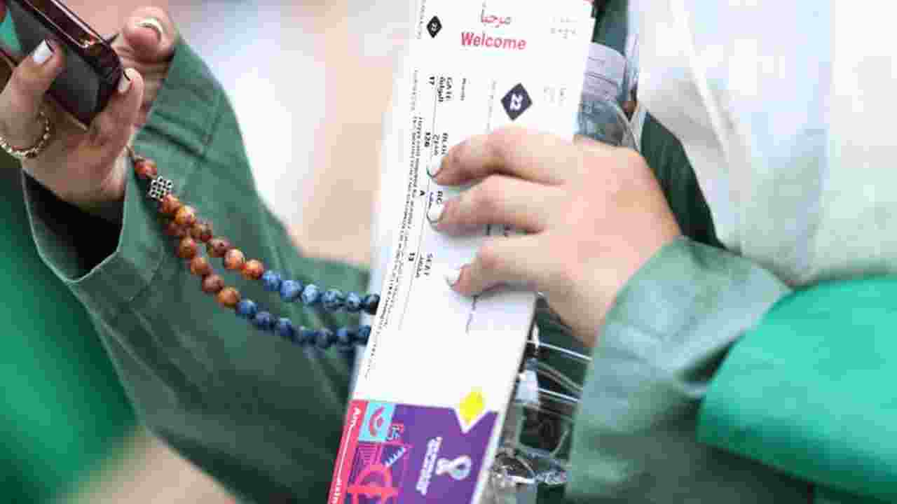 Close-up of a woman in a green dress wearing a white head covering, checking her phone with her right hand while holding a FIFA World Cup Qatar 2022 match ticket in her left hand. Beads extend from her right hand to behind her left.