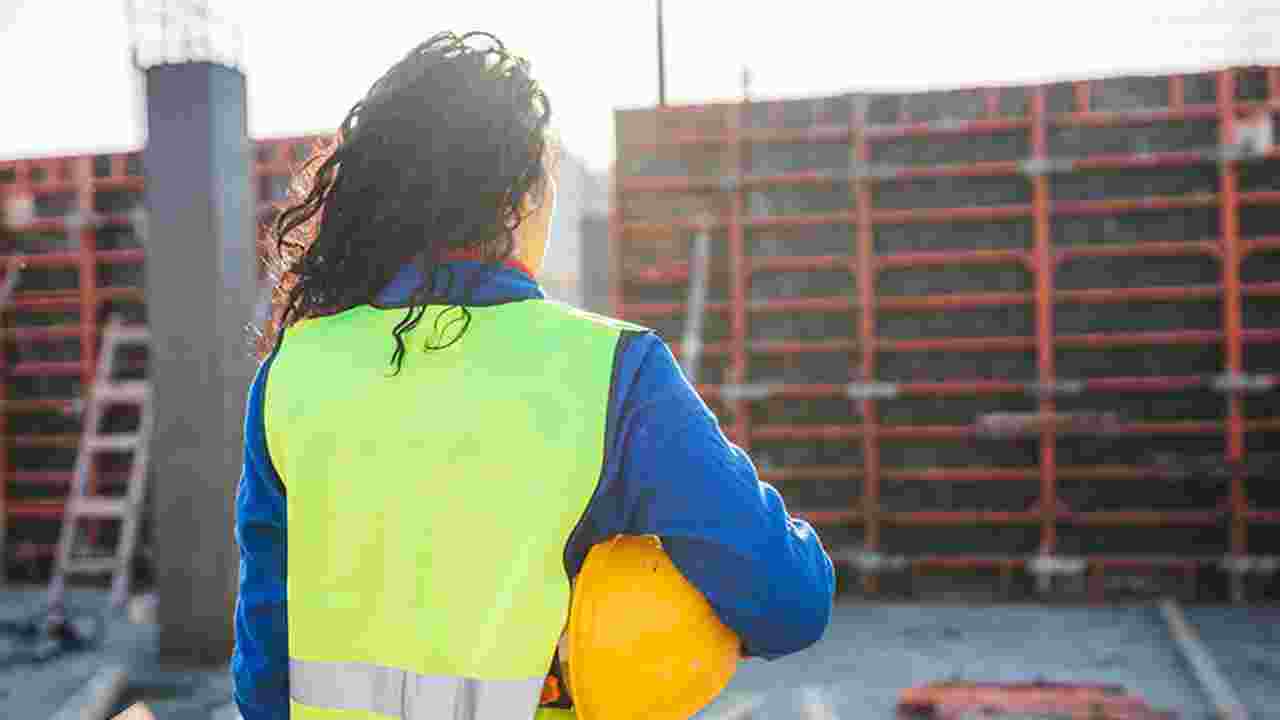 A female worker in a safety vest faces away from the camera, looking at a construction site, with a safety helmet tucked under her arm. 